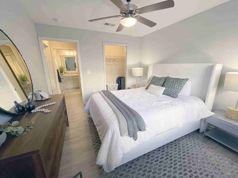 Greenville, SC, Luxury Apartments Near Libery Bridge - The Aventine Greenville - A Bedroom With A White Upholstered Bed, Grey Pillows, And A View Into A Walk-In Closet And En-Suite Bathroom.