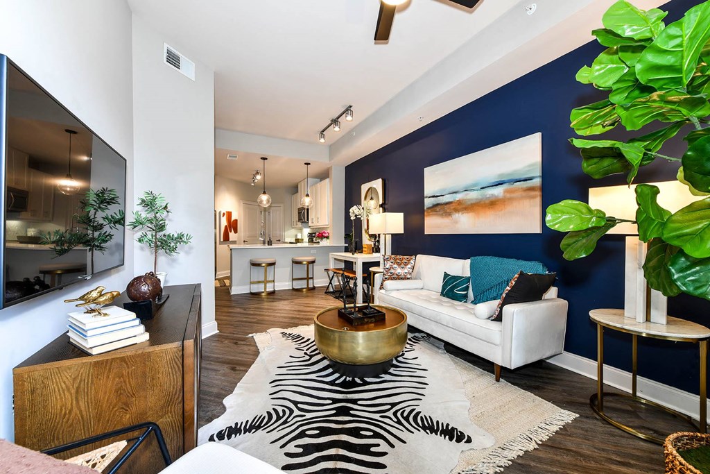Buckhead, GA, Luxury Apartments Near Phipps Plaza- 92 West Paces - Furnished Living Room with Hardwood Flooring, Area Rug, Blue Accent Wall, White Sofa, Coffee Table, Side Tables with Lamps, Wooden Cabinet, Wall-Mounted TV, Houseplants, and View of Kitchen.
