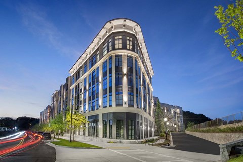 A modern building with a curved facade and large windows is illuminated from within, with a clear blue sky above and a road with moving cars in the foreground.