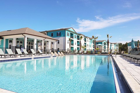 Luxury Apartments in Flagler Beach, FL, with In-Unit Washer and Dryer - Preserve at Flagler Beach - Resort Style Pool With Lounge Chairs, Palm Trees, Clubhouse, And Apartment Buildings.
