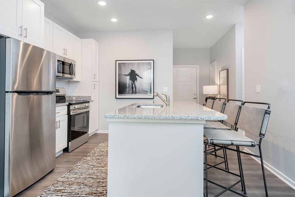 One-Bedroom Apartments in Wyoming, MI - The Retreat - Kitchen with Wood-Style Vinyl Plank Flooring, White Shaker-Style Cabinetry, Granite Countertops, Recessed Lighting, Stainless-Steel Energy-Efficient Appliances, Eat-in Kitchen Island with Breakfast Bar, 4 Bar Stools, Long Rug, Large Wall Arts, Lamp, and a View of the Doorway.