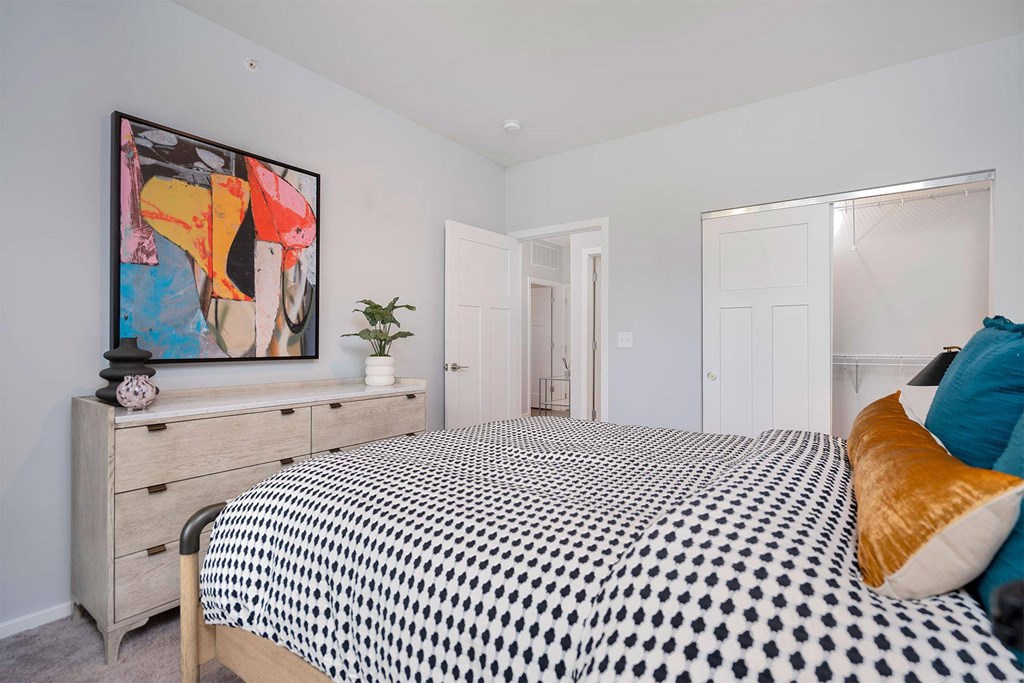 Apartments in Wyoming, MI, with In-Unit Washer and Dryer - The Retreat - Bedroom with Carpet Flooring, Bed, Wooden Dresser, Large Wall Art, Home Decor, Oversized Closet with Sliding Doors, and an Opened Bedroom Door.