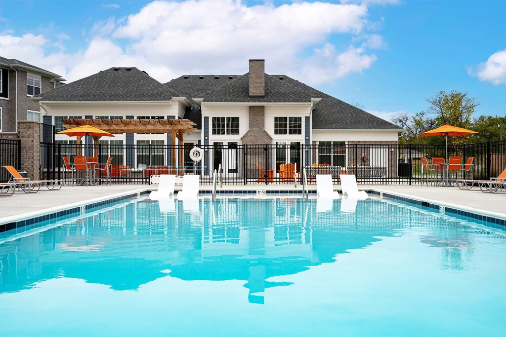 2-Bedroom Apartments in Wyoming, MI - The Retreat - Outdoor Heated Swimming Pool with In-Water Loungers, Surrounded by Orange Loungers, Poolside Seating with Umbrellas, and Fence, Overlooking the Clubhouse and Apartments.