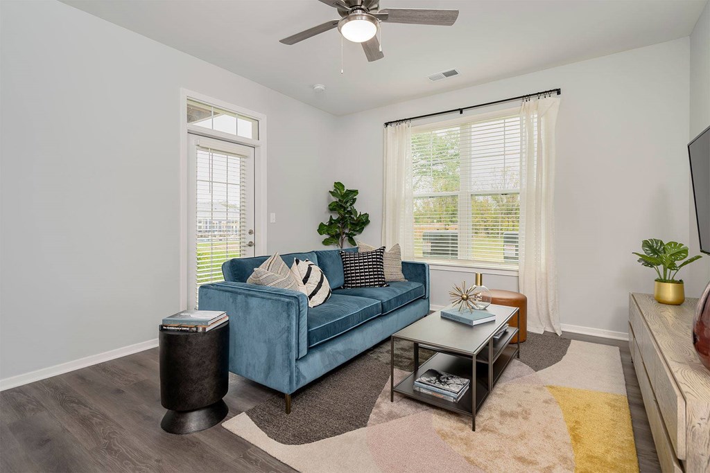 Wyoming, MI, Apartments with In-Unit Washer and Dryer - The Retreat - Living Room with Wood-Style Vinyl Plank Flooring, Large Rug, Couch, Coffee Table, Black Cylindrical Side Table, Home Decor, Plants, Tan Stool, Media Console Cabinet, Mounted Flat Screen TV, Ceiling Fan, Glass Door with Blinds, and Large Window with Floor Length Curtains and Blinds.