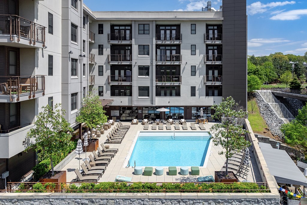 A large swimming pool is surrounded by lounge chairs and umbrellas in front of a multi-story apartment building.