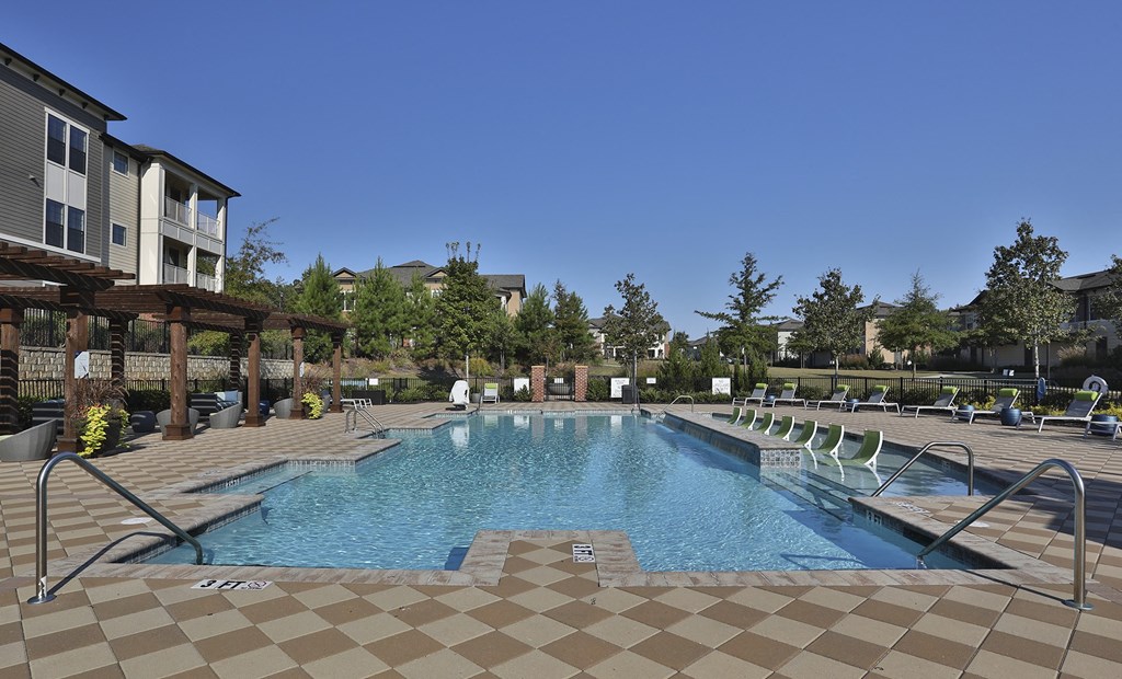 Greenville, SC Luxury Apartments - Velo Verdae - Resort-Style Swimming Pool with Sundeck, Submerged and Poolside Lounge Chairs, Seating Areas Under Pergolas, Checkered Tiles, and View of Apartment Building and Surrounding Trees