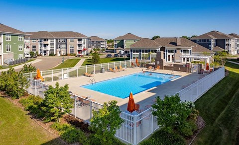 Kalamazoo, MI, Apartments for Rent - Parkway Flats - Aerial View of Heated Outdoor Saltwater Pool with Sun Deck, Surrounded by White Fence, Orange Loungers and Umbrellas, Overlooking the Landscaping, Clubhouse, and Apartments.