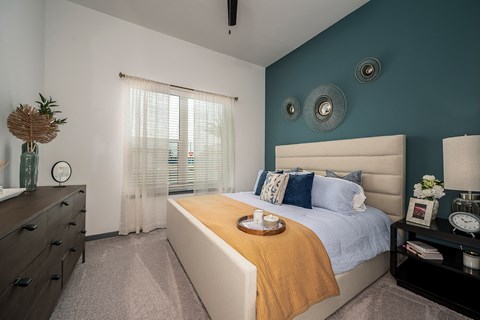 Apollo Beach, FL, Luxury Apartments Near Anna Maria Island - The Citizen at Waterset - Bedroom With Upholstered Bed, Teal Accent Wall, Nightstand, Lamp, Dresser, and Large Window With Coverings.