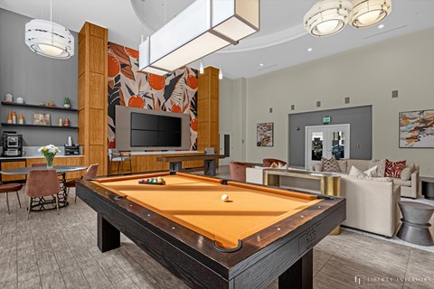 Dog-Friendly Luxury Apartments in Orlando, FL - NORA - Resident Club Room with Pool Table, Sofas, Armchairs, Kitchen with Dining Table and Chairs, High Ceiling, and Bright Lighting.