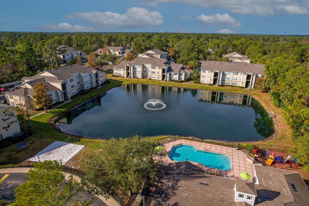 A large swimming pool surrounded by a fence and a small lake in the middle of a housing complex.