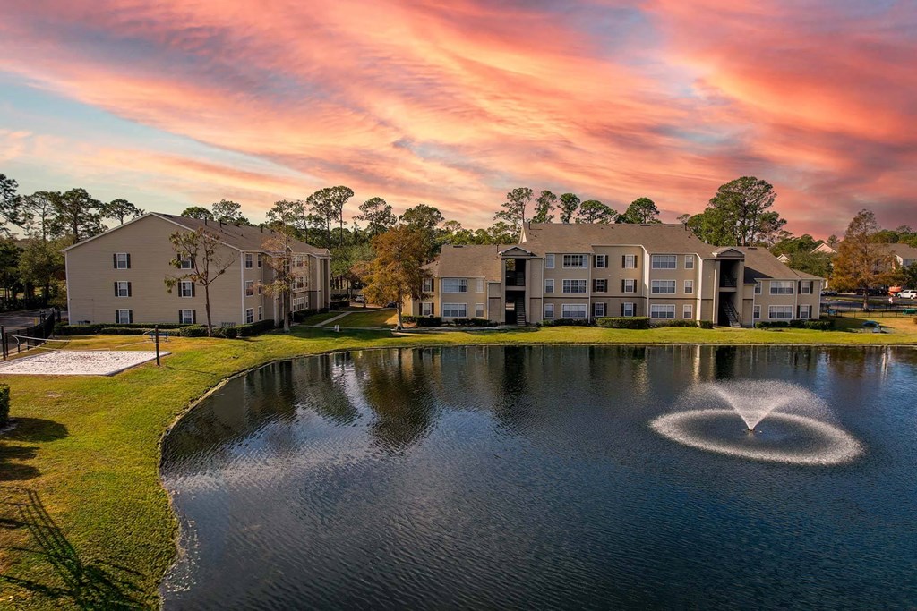 A sunset view of a lake with a fountain in the middle and apartment buildings in the background.