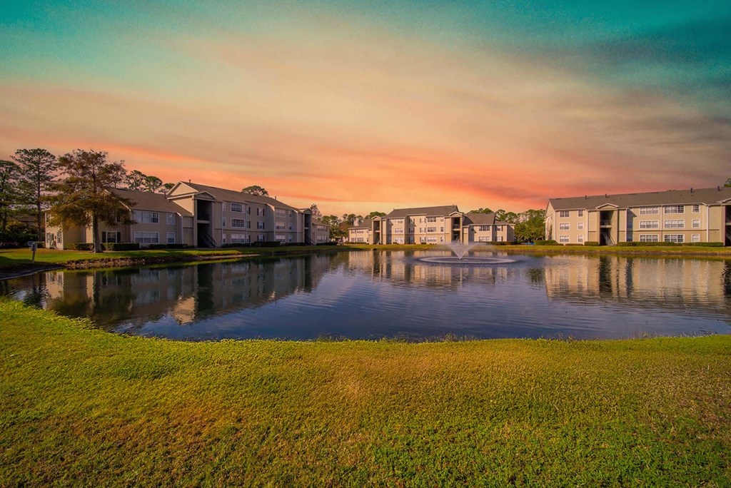 A serene landscape with a lake, grass, and buildings in the background.