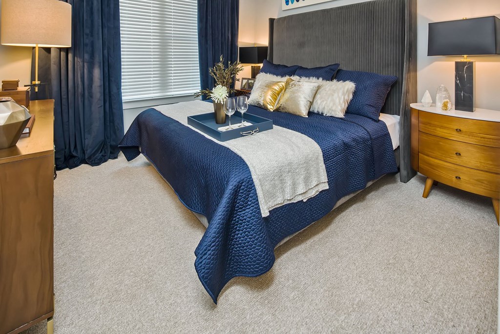 1 BR Apartments in Tampa FL - Society Westshore - Furnished Bedroom with Carpeted Floor, Bed, Wooden Nightstands with Grey Lamps, Wooden Dresser, Standing Lamp, and Large Window with Blinds and Curtains