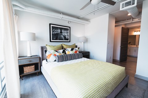 Apartments for Rent in Midtown Memphis, TN - The Citizen - Bedroom With Large Bed, Ceiling Fan, Nightstands, Lamps, Wall Art, Large Windows, and Bathroom Entry.
