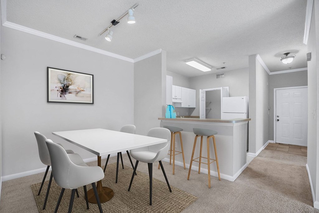 A white dining table with four chairs and a kitchen area in the background.