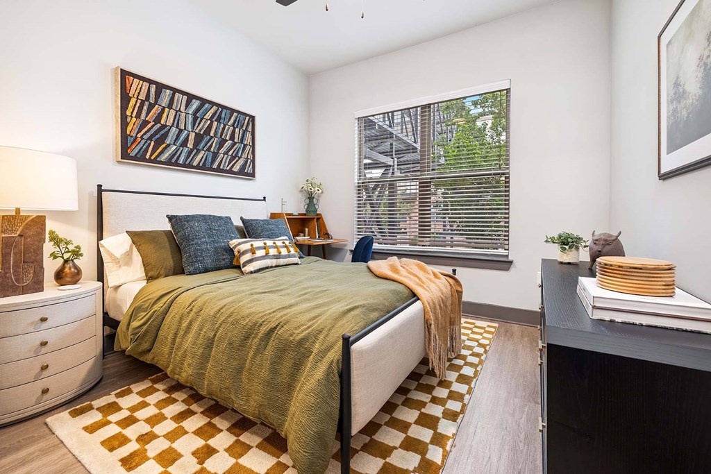 A bedroom with a bed, a dresser, a window with blinds, a painting on the wall, a plant on the nightstand, and a rug on the floor.