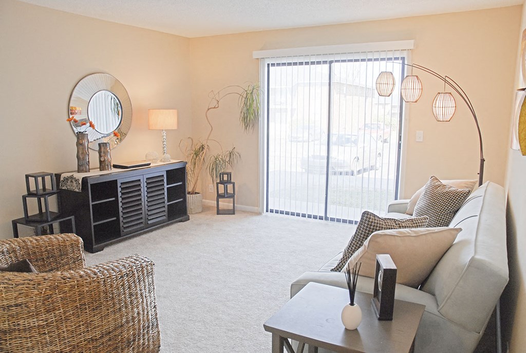 2-Bedroom Apartments in Smyrna, TN - The Grove at Enon Springs - Living Room with Carpeted Flooring, Couch, Wicker Barrel Chair, Side Table, Storage Cabinet, Floor Lamp, and Sliding Glass Door.