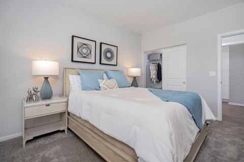 Pet-Friendly Apartments in Middletown, OH - Echo Ridge Apartments - Modern Bedroom With Wood Bed Frame, White Bedding, Blue Accent Pillows, Bedside Lamps, Soft Carpet Flooring, And Open Closet.