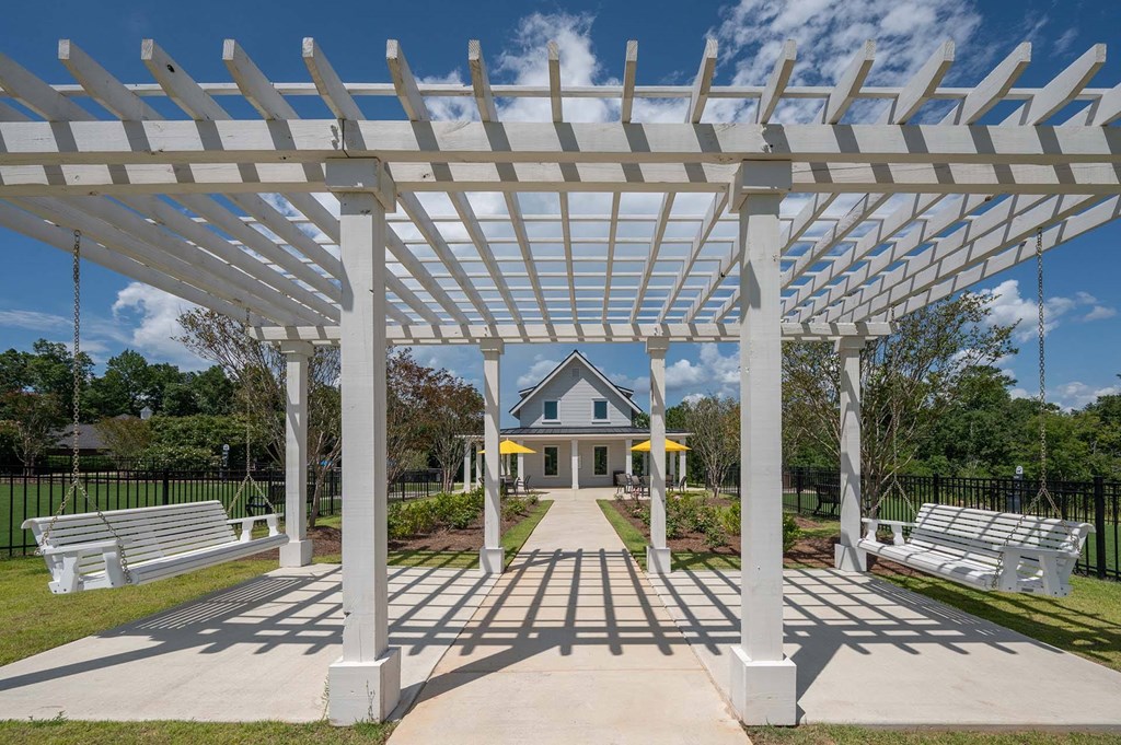 A white pergola structure is in the foreground of a park with benches and a building in the background.
