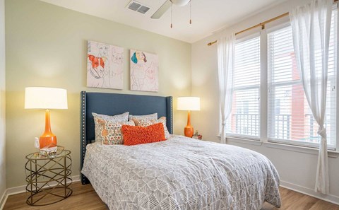 Pet-Friendly Apartments in Tampa, FL - Havana Square - Bedroom with Vinyl Plank Flooring, Bed, Nightstands, Lamps, Wall Paintings, Ceiling Fan, and Large Windows with Coverings.