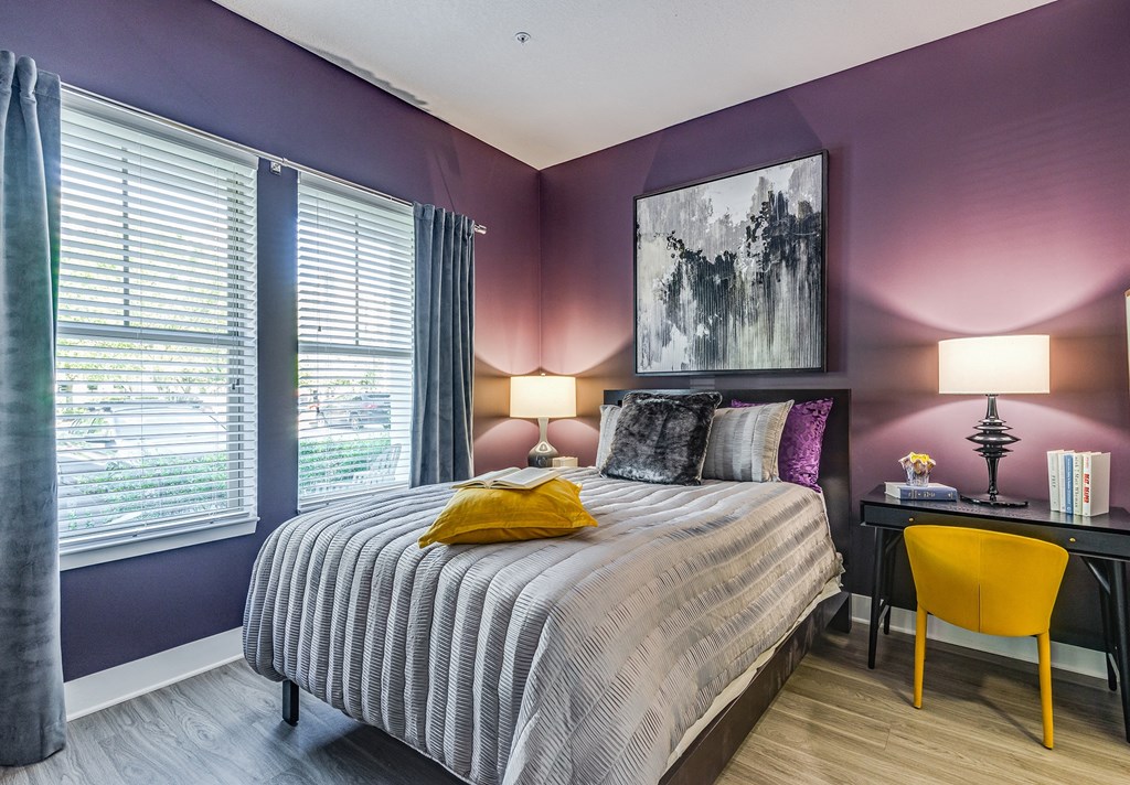 1 BR Luxury Apartments in Altamonte Springs FL - Nine12 Gateway - Furnished Bedroom with Wood-Style Vinyl Plank Flooring, Bed, Nightstand with Lamp, Desk with Yellow Chair and Lamp, Two Windows with Curtains and Blinds, Purple Walls, and Framed Art