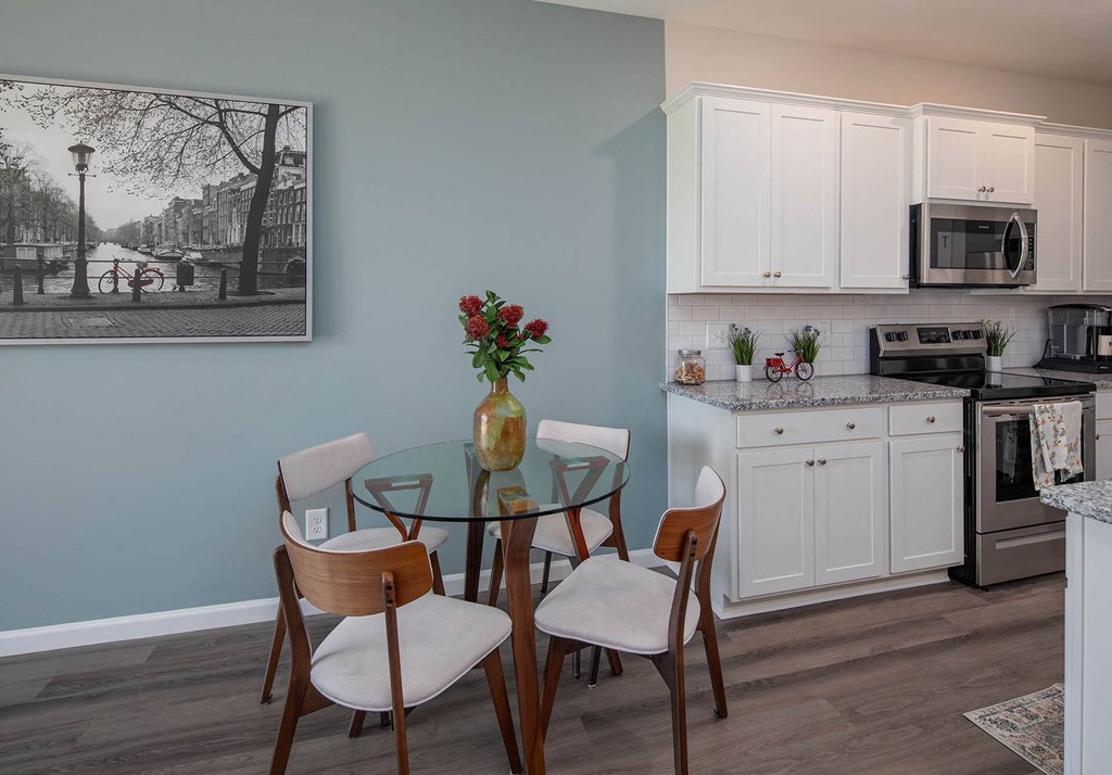 a dining area with a glass table and four chairs and a kitchen in the background