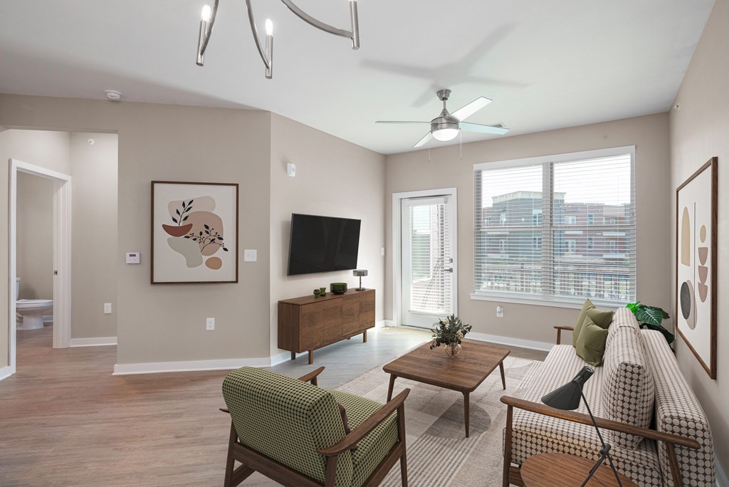 Mt. Juliet, TN Apartments Near Percy Priest Lake - The Enclave at Providence - Living Room with Hardwood-Style Flooring, Area Rug, Sofa, Armchair, Coffee Table, Media Cabinet, Wall-Mounted TV, Ceiling Fan, Large Window, and Glass Door with Access to Balcony.