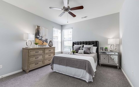 One-Bedroom Apartments in Kalamazoo, MI - Parkway Flats - Bedroom with High Ceilings, Ceiling Fan, Bed, Dresser, Nightstand, Lamps, Decorative Plants, Large Wall Art, Carpet Flooring, and 2 Corner Windows with Wood-Style Blinds.