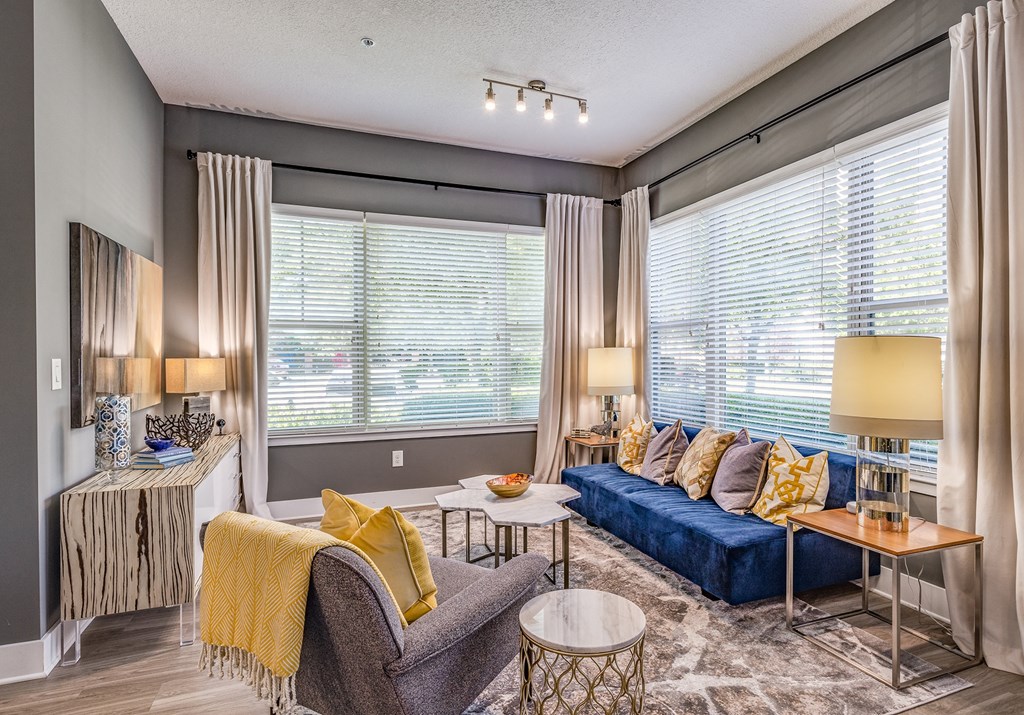 Altamonte Springs, FL Luxury Apartments - Nine12 Gateway - Furnished Living Room with Wood-Style Vinyl Plank Flooring, Area Rug, Sofa, Armchair, Cofeetable, Side Tables with Lamps, Storage Dresser with Wood-Style Design, and Two Large Windows with Blinds and Curtains
