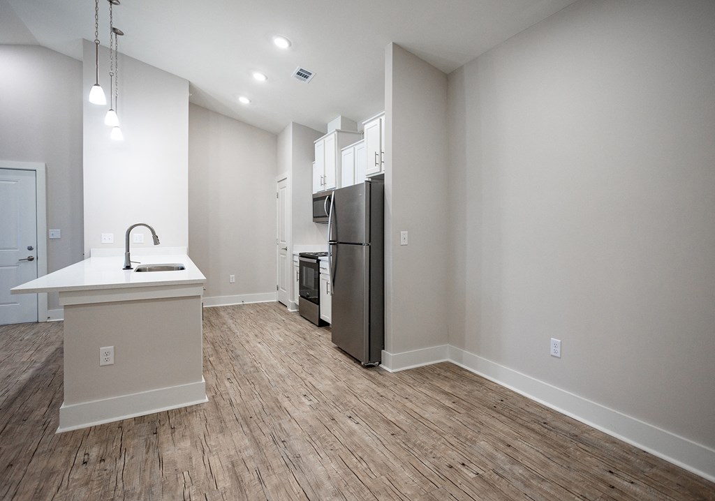 an empty living room with a kitchen with a refrigerator and a sink