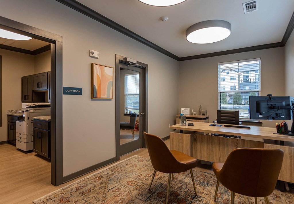 a room with a desk and chairs and a kitchen in the background