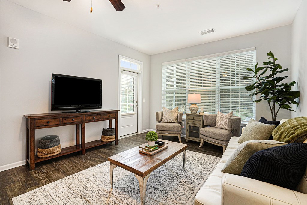 Grand Rapids, MI, Apartments With In-Unit Washer and Dryer - Knapp’s Corner Flats - Living Room with Wood-Style Vinyl Plank Flooring, 9-Foot Ceiling, Ceiling Fan, Console Table, Flat-Screen TV, Storage Baskets, Couch, Armchairs, Coffee Table, Side Table, Large Rug, Decorative Plants, and Large Window with Blinds.