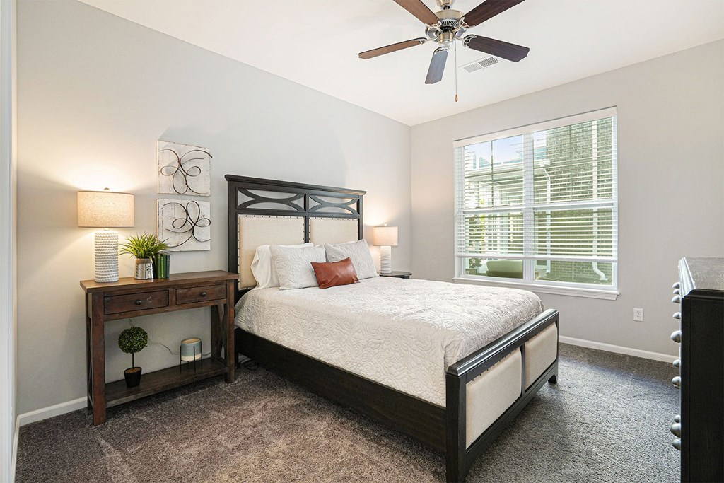 Three-Bedroom Apartments in Grand Rapids, MI - Knapp’s Corner Flats - Bedroom with Carpet Flooring, 9-Foot Ceiling, Ceiling Fan, Queen Size Bed, Nightstands, Lamps, Wall Arts, Decorative Plants, Dresser, and a Large Window with Blinds.