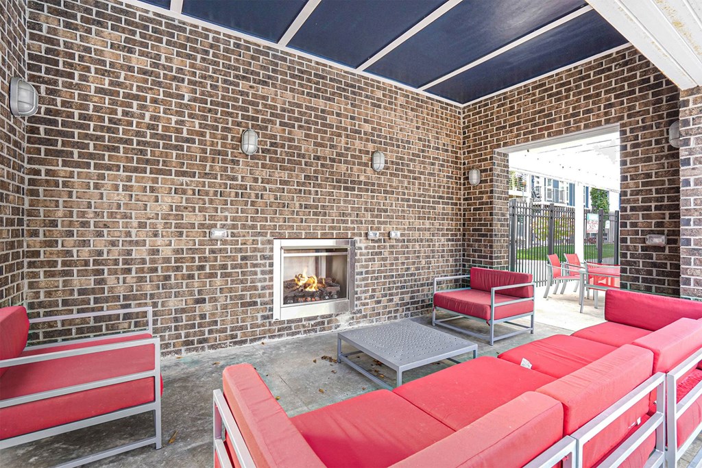 3-Bedroom Apartments in Grand Rapids, MI - Knapp’s Corner Flats - Outdoor Lounge Featuring a Brick Wall with an Integrated Fireplace and Light Fixtures, Furnished with a Red Couch and Armchairs, a Low Coffee Table, and a View of the Outdoor Seating Area.