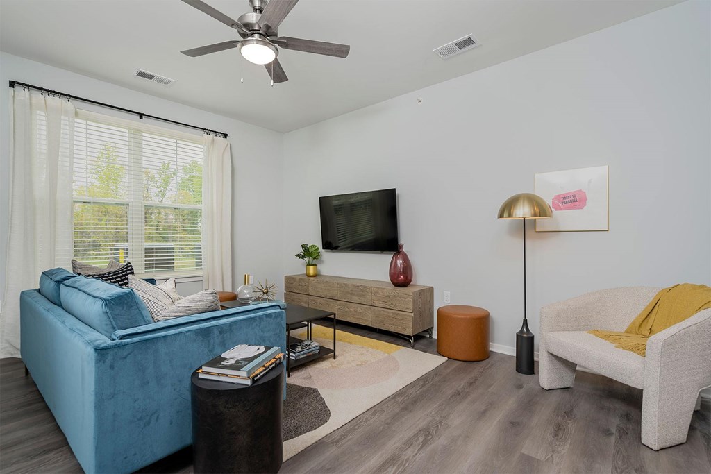One Bedroom Luxury Apartments In Jeffersonville, IN - The Laurel - Living Area With Wood-Style Floor, Large Window, Sofa, Armchair, Coffee Table, Wall-Mounted TV, And Ceiling Fan.