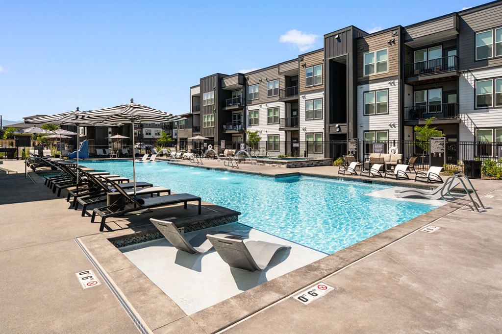 A swimming pool surrounded by lounge chairs and umbrellas in front of apartment buildings.