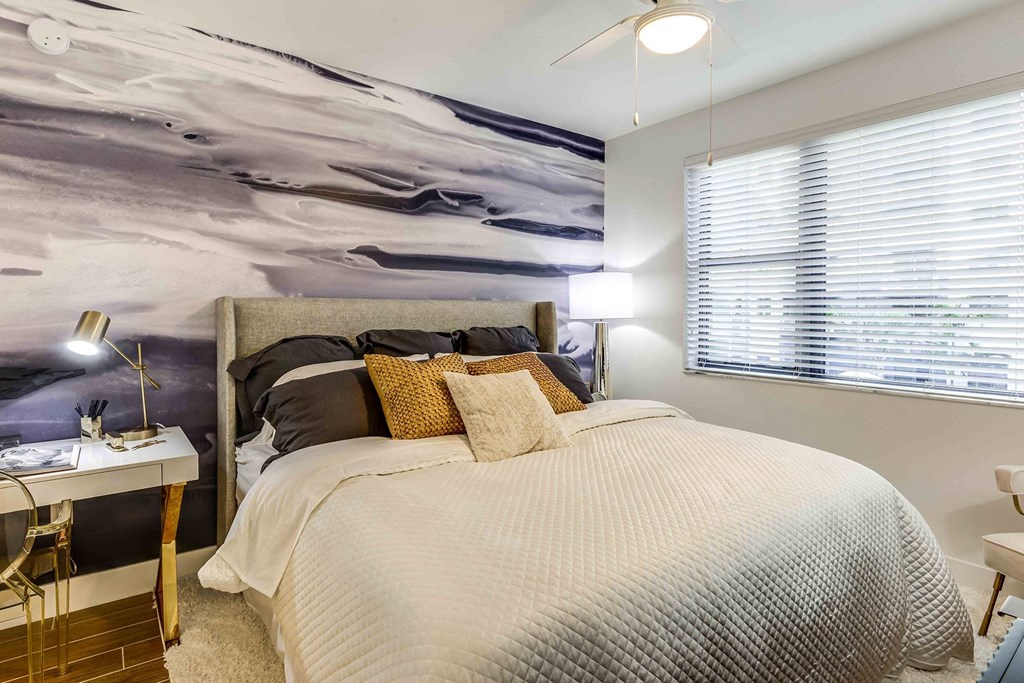 Luxury Apartments in North Miami Beach, FL with In-Unit Washer and Dryer - Lazul - Bedroom with Porcelain Wood-Style Plank Flooring, Plush Area Rug, Large Bed, Desk with Lamp, Wall Mural, and Large Window.