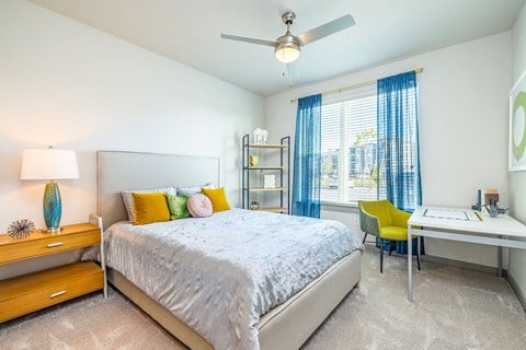 Pet-Friendly Apartments In Daytona Beach, FL - Madison Pointe - Bedroom With Bookshelf, Desk, Chair, Nightstand, Bed, Ceiling Fan, And Large Window.