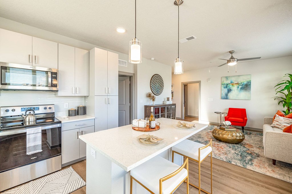 Pet Friendly Apartments in Daytona Beach, FL - Madison Pointe - Designer Kitchen With Modern Cabinetry, Stainless Steel Appliances, Wood-Style Plank Flooring, Breakfast Bar with Bar-Style Chairs, Quartz Countertops, Designer Pendant Lighting, and View of Living Room