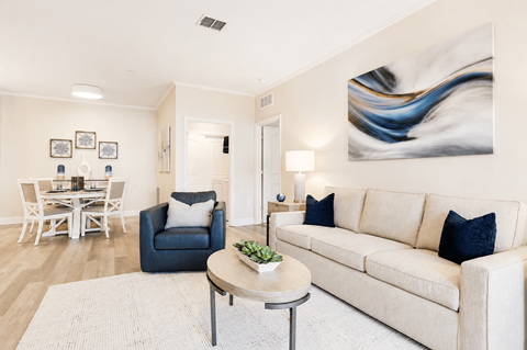 A living room with a white couch, a blue chair, a dining table with chairs, a lamp, and a painting on the wall.