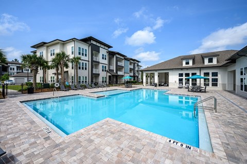 1-Bedroom Apartments in Pensacola, FL - Preserve at Perdido - Resort-Style Swimming Pool Surrounded by Paver Deck, Furnished with Lounge Chairs, Tables with Umbrellas, and Apartment Building Backdrop.