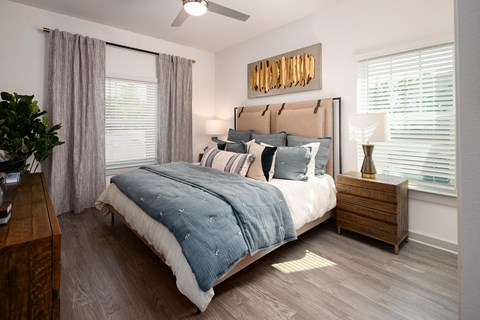 Pet-Friendly Luxury Apartments in Ridgeville, SC - Preserve at Ridgeville - Bedroom With Upholstered Bed, Blue Bedding, Wooden Nightstand With Lamp, Two Large Windows With Curtains And Blinds.