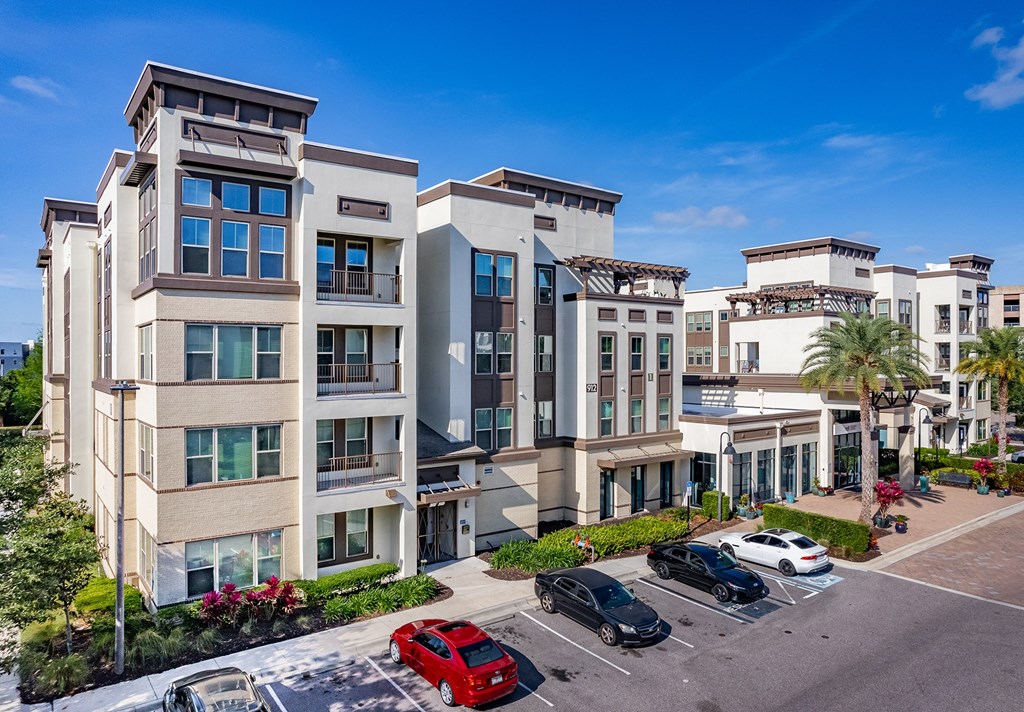 Luxury Apartments in Altamonte Springs, FL - Nine12 Gateway - Tan and White Property Exterior, with Street View of Parking, Landscaped Bushes, Plants, and Palm Trees