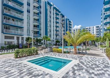 A swimming pool in the middle of a tiled area surrounded by palm trees and bushes.