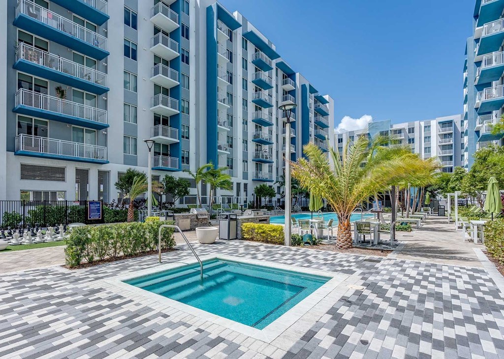 A swimming pool in the middle of a tiled area surrounded by palm trees and bushes.