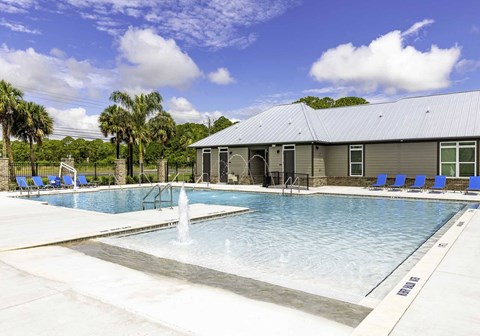 One-Bedroom Apartments in Titusville, FL - Riverwalk Grand - Resort Style Pool With Shallow Water Lounge Area, Splash Fountain, Blue Chairs, Palm Trees, And Clubhouse Building.