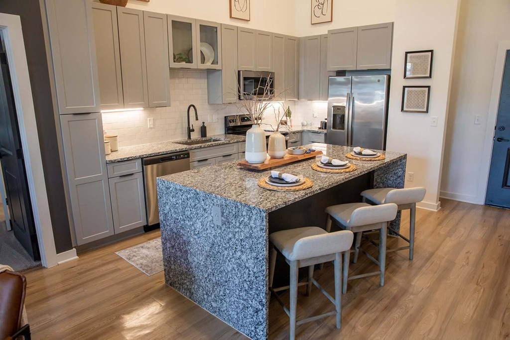 Upscale Apartments in Nashville TN - Sterling Nashville West - Modern Kitchen with Hardwood-Style Flooring, Large Waterfall Cooking Island, Granite Countertops, Breakfast Bar Stool Seating, Grey Cabinetry, Stainless Steel Appliances, Underglow Lighting, and White Tile Backsplash