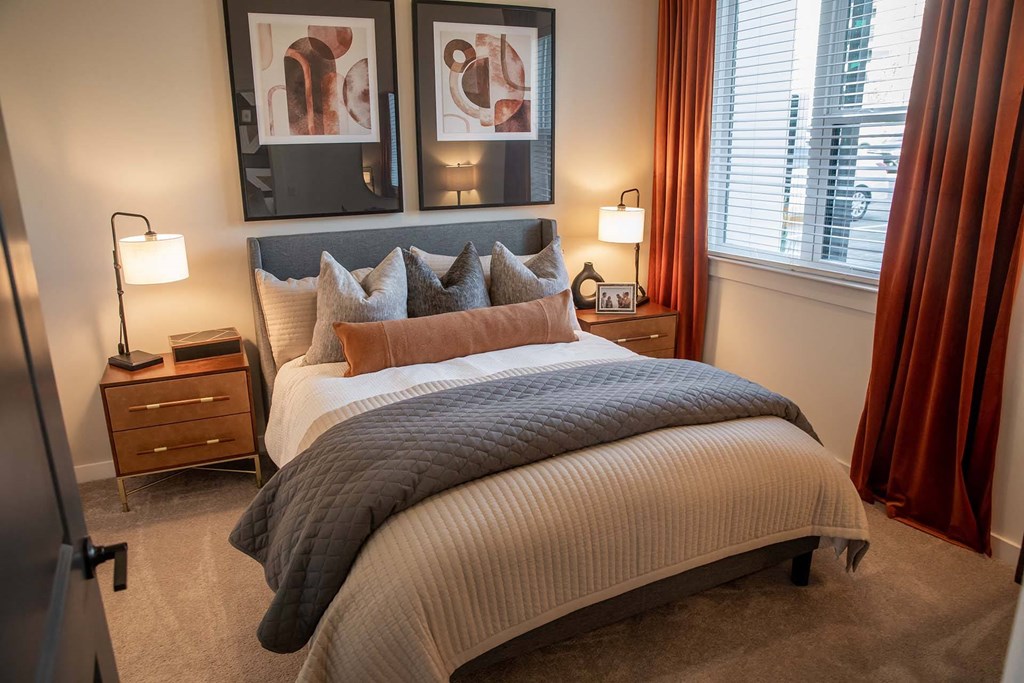 One BR Luxury Apartments in Nashville TN - Sterling Nashville West - Furnished Bedroom with Carpeted Floor, Large Bed, Wooden Nightstands with Lamps, Framed Wall Art, Large Windows with Blinds, and Orange Curtains