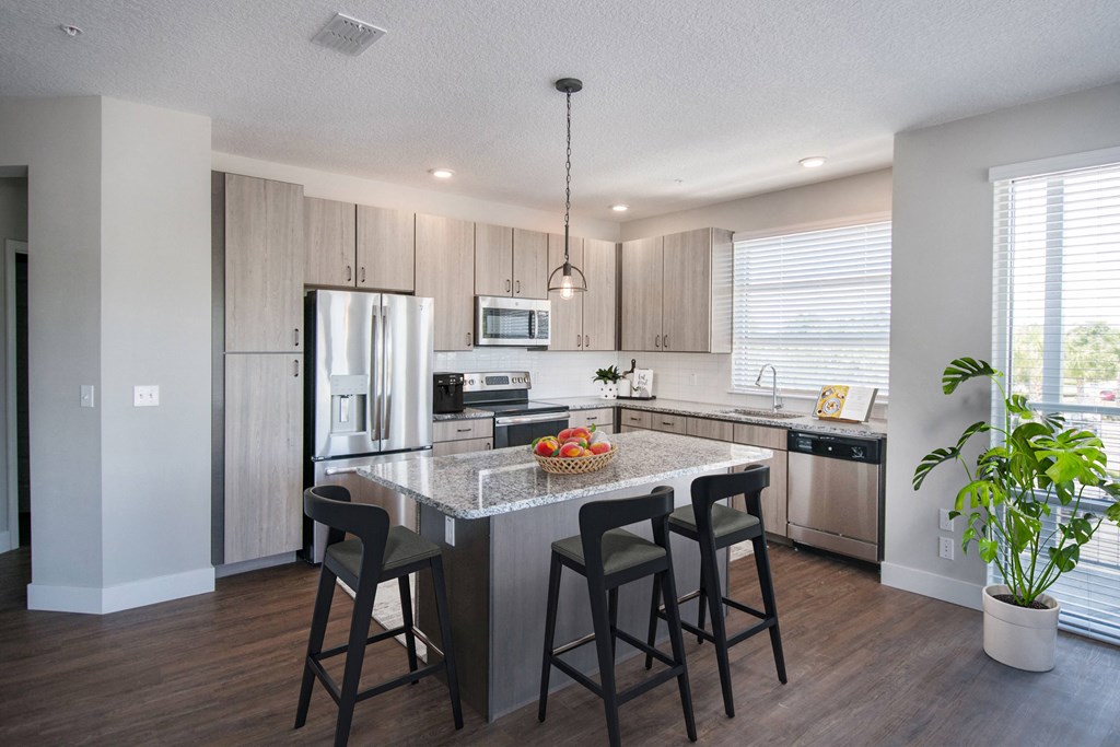 One-Bedroom Apartments in Maitland - The Parker at Maitland Station - Kitchen with Wood Vinyl Plank Flooring, Tan Flat-Panel Cabinetry, Granite Countertops, Stainless Steel Appliances, Kitchen Island with Seating, and Pendant Light.