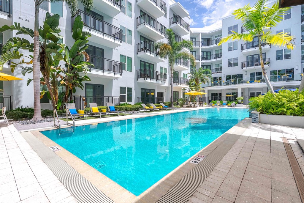 Tampa, FL Apartments Near Tampa Riverwalk - Society Westshore - Swimming Pool with Poolside Lounge Seats, Umbrella Tables and Chairs, Palm Trees, and View of Apartment Building.